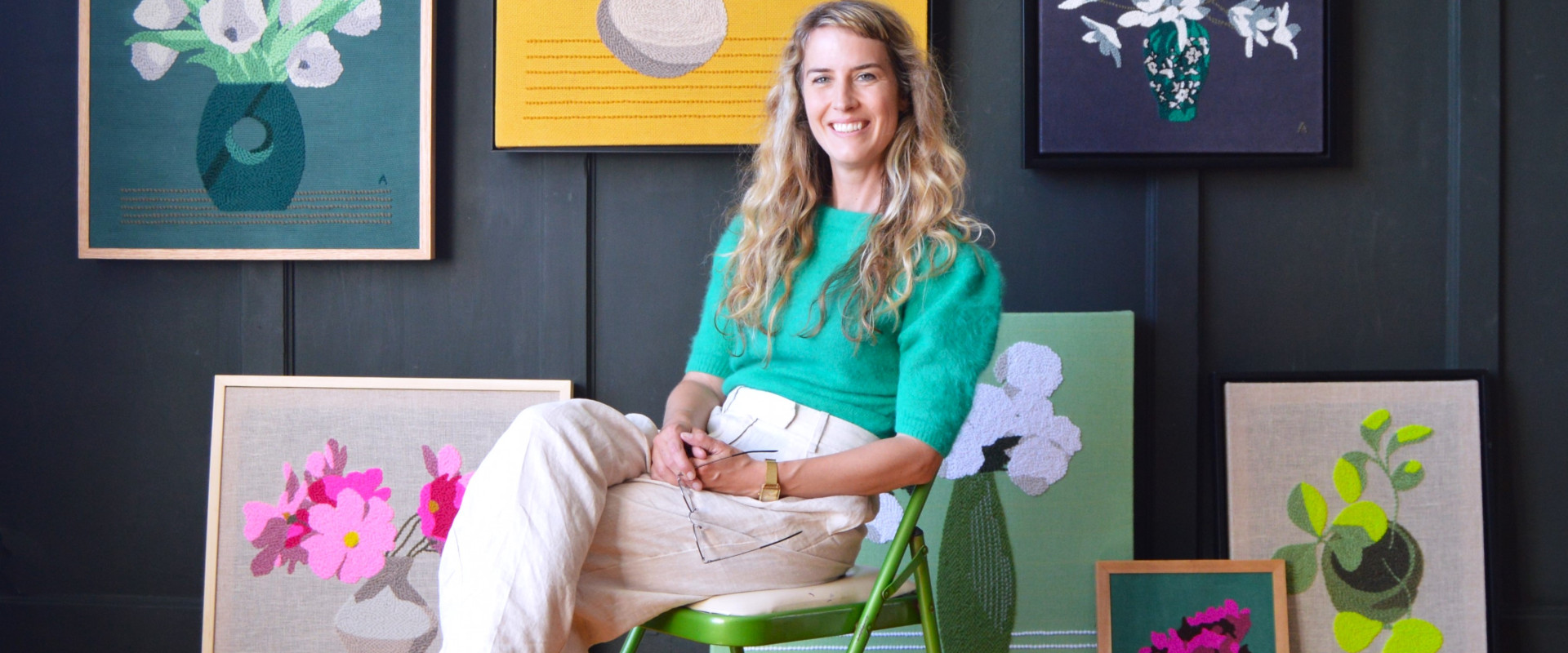 A blonde woman in a green shirt and beige pants sits in a green chair in front of 7 hooked rug art pieces of flowers in vases, in a variety of colours of flowers and vaes, on coloured backgrounds