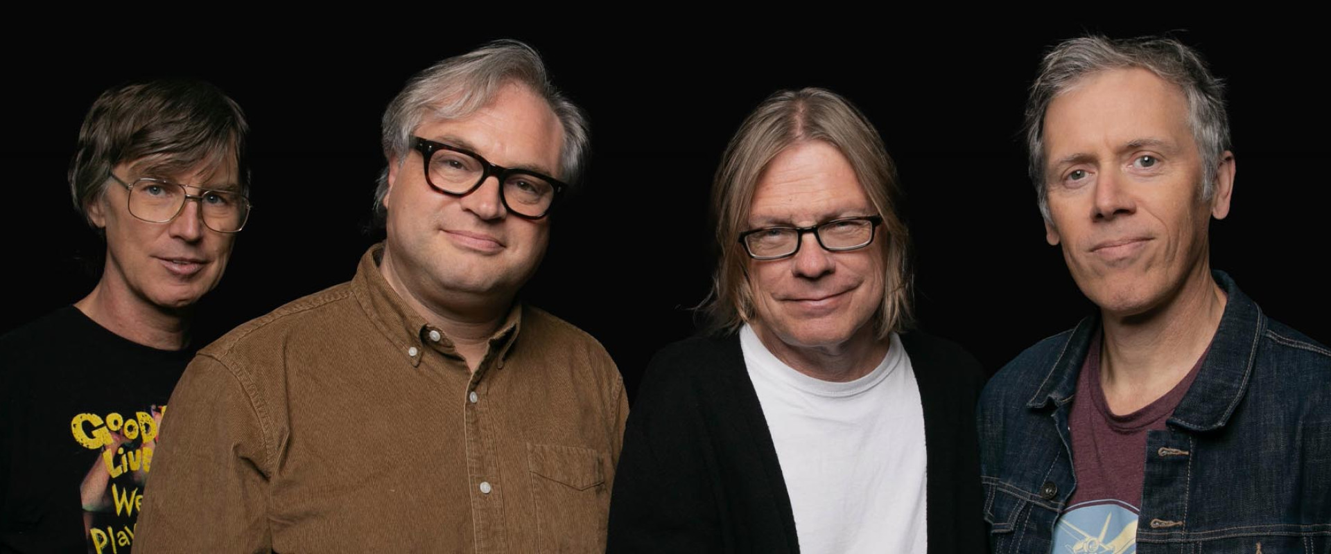 The four members of Trans-Canada Highwaymen are looking at the camera with smiles on their faces against a black backdrop. They are all wearing casual clothes.