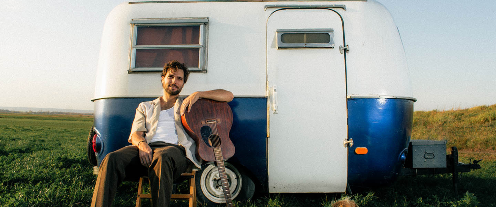 Picture of Daniel James McFadyen with a guitar and dog in front of a trailer. 