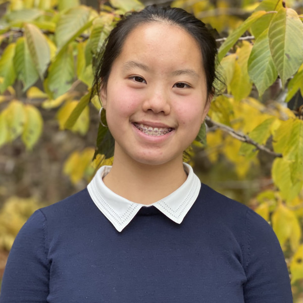 Girl in navy sweater and white collared shirt standing in front of leaves